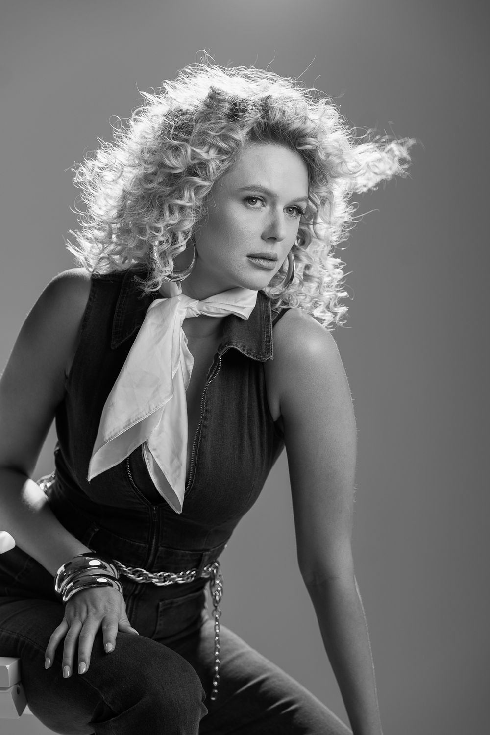Black and white studio portrait showing curly blonde backlit hair, black outfit with white necktie, and chain belt.