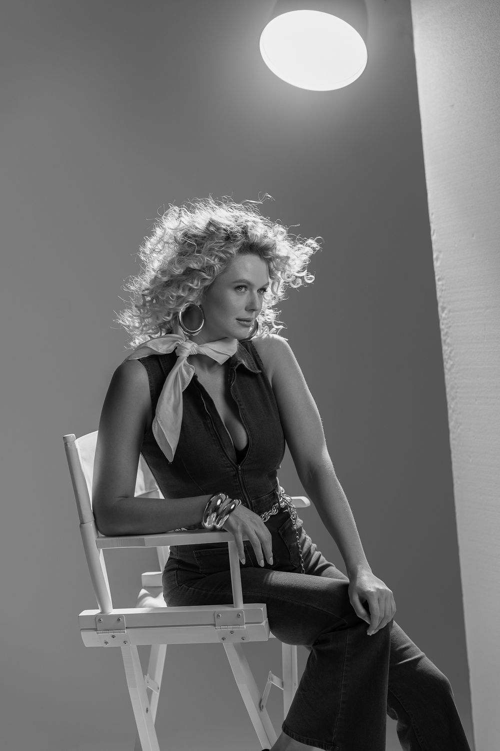 A black and white fashion photo shows a figure in a dark sleeveless outfit seated on a director's chair under dramatic lighting.