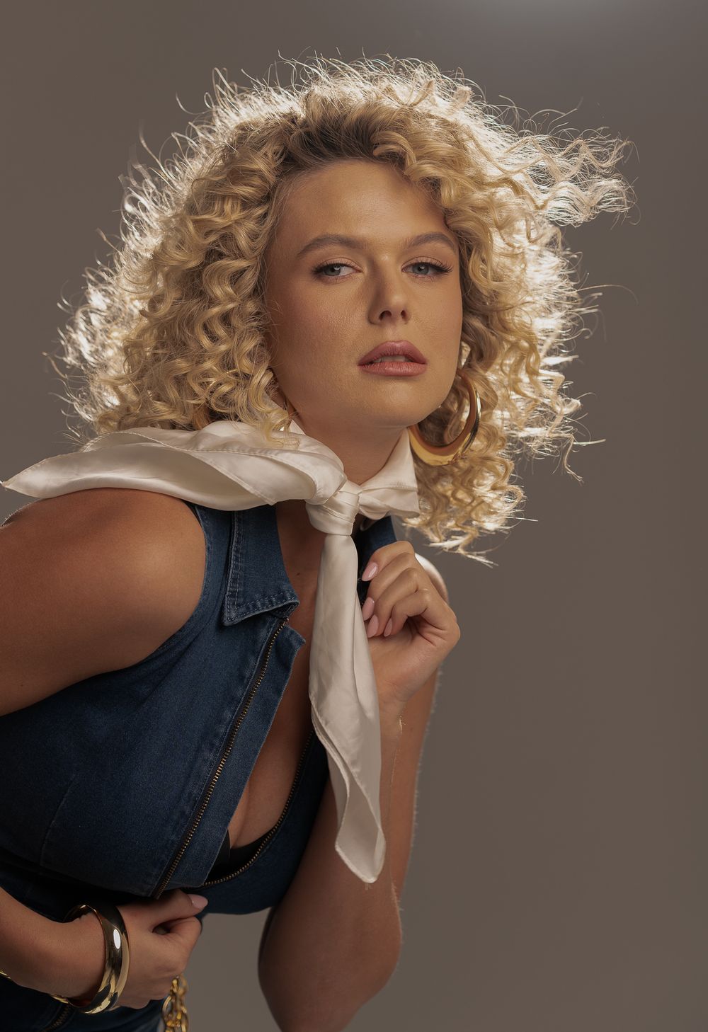 Studio portrait with curly blonde hair and white scarf against dark background wearing denim dress.