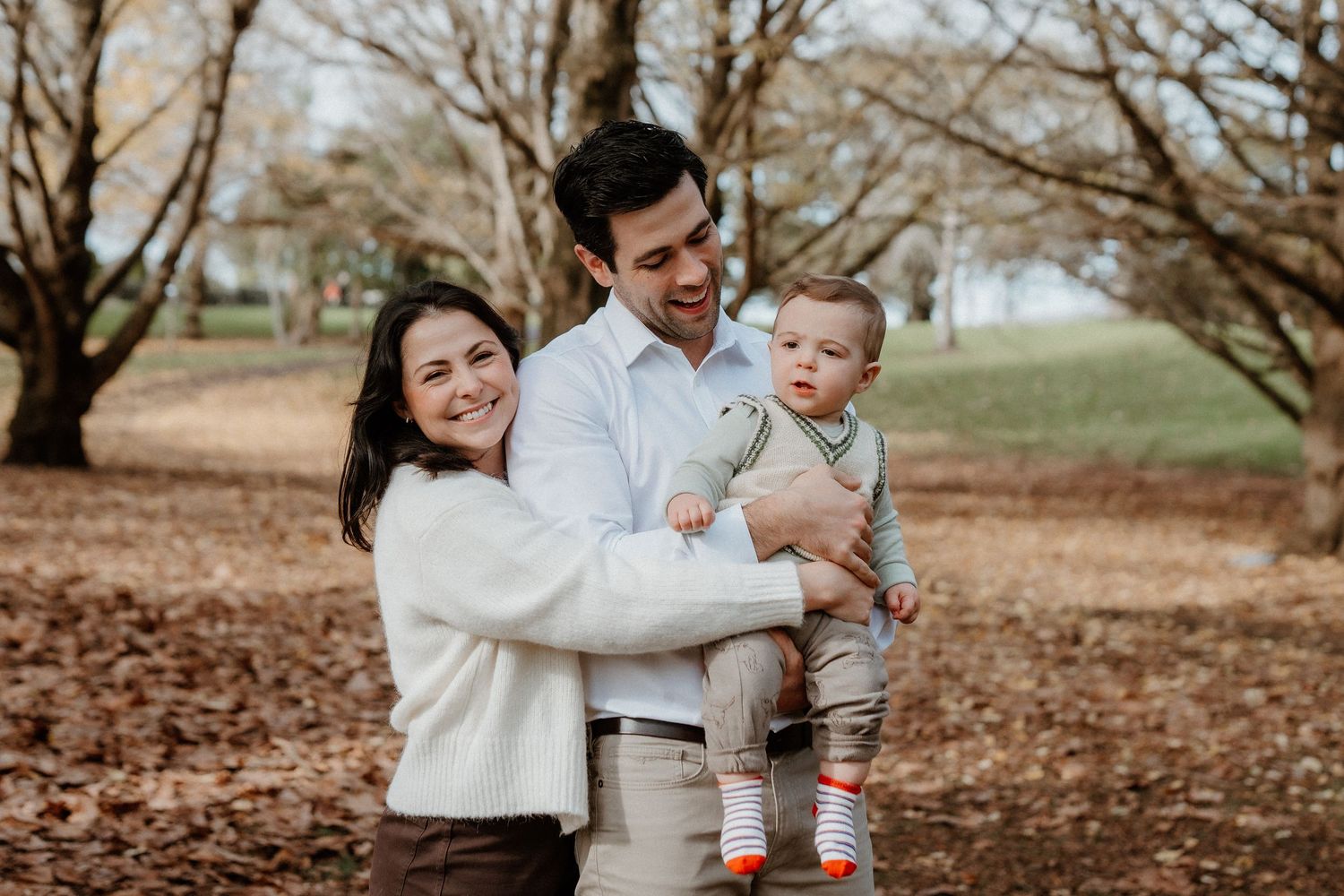A family enjoys a tender moment together on an autumn day with fallen leaves and bare trees in the background.