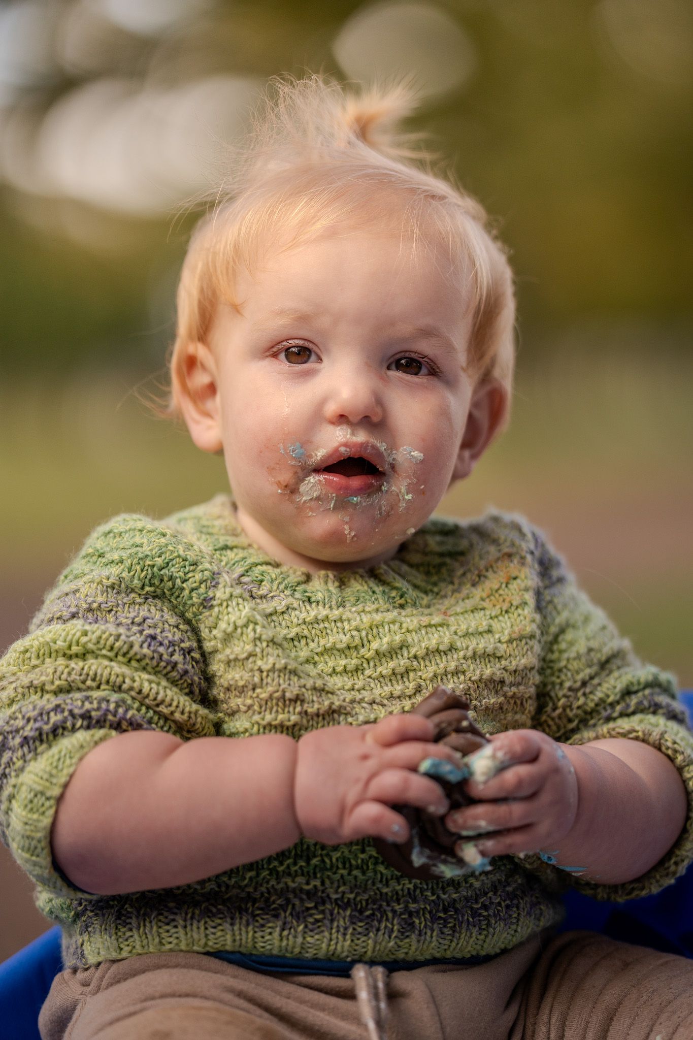 A toddler in a green knit sweater has chocolate smeared around their mouth while holding something outdoors.
