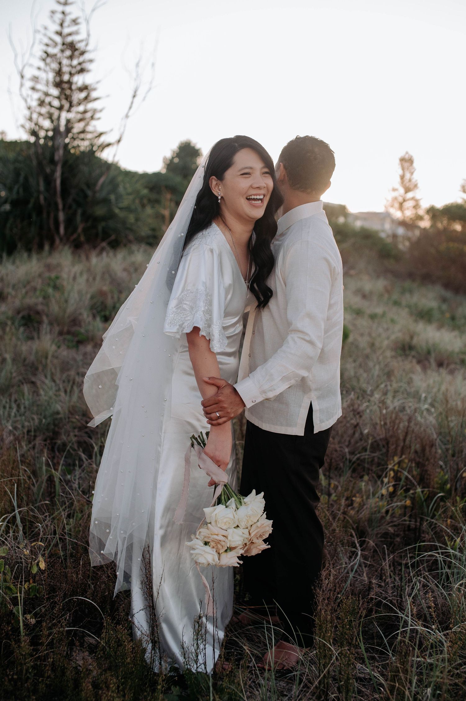 A couple in wedding attire shares a joyful moment in a rustic outdoor setting at sunset.