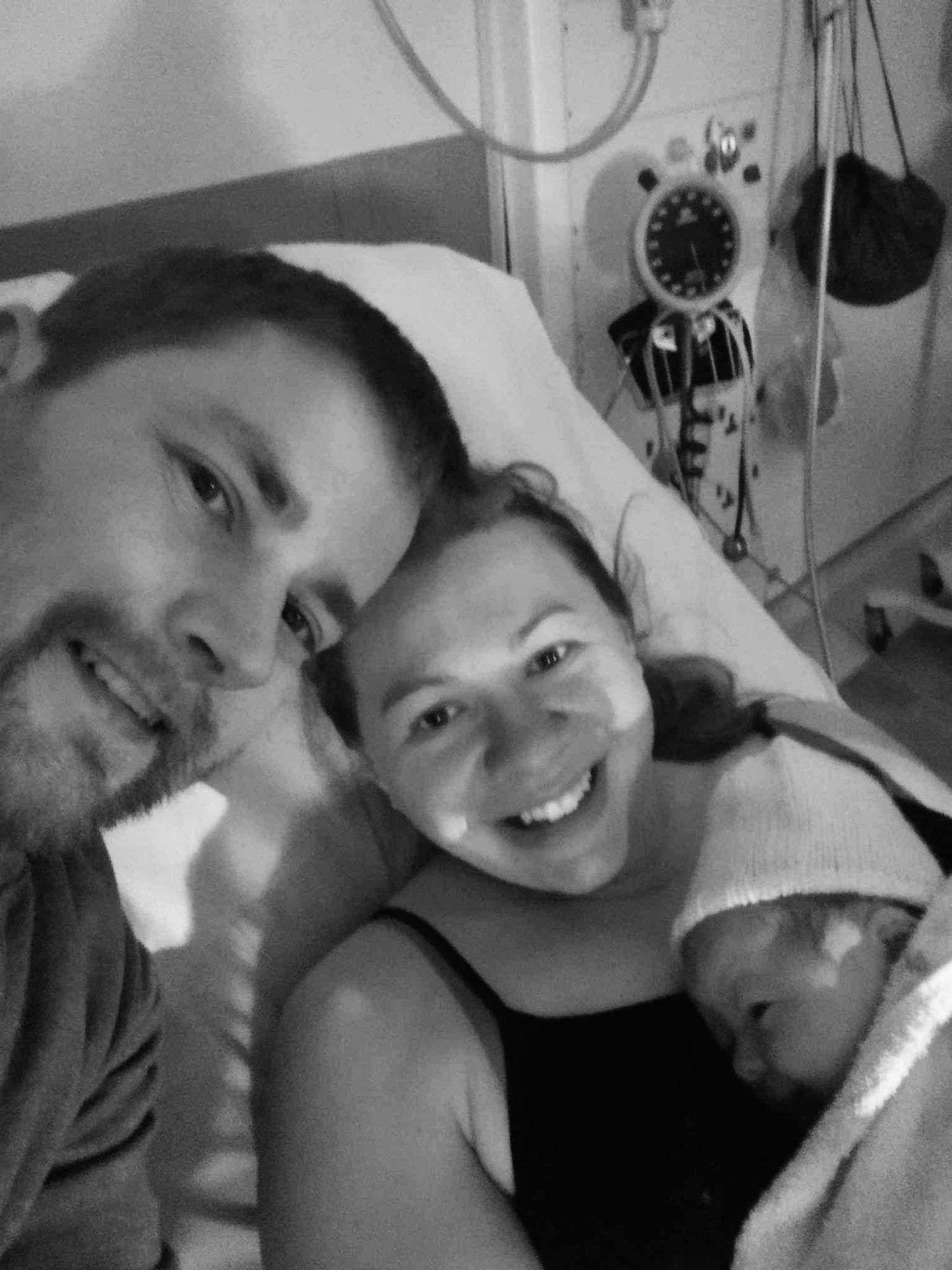 A black and white photo shows a joyful moment between parent and newborn baby resting together.