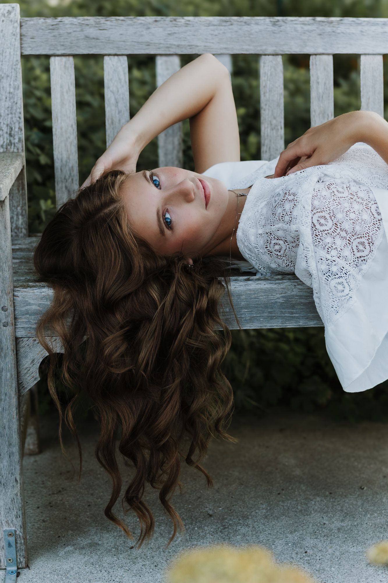 Young woman in white dress poses on gravel path with flowers in background.