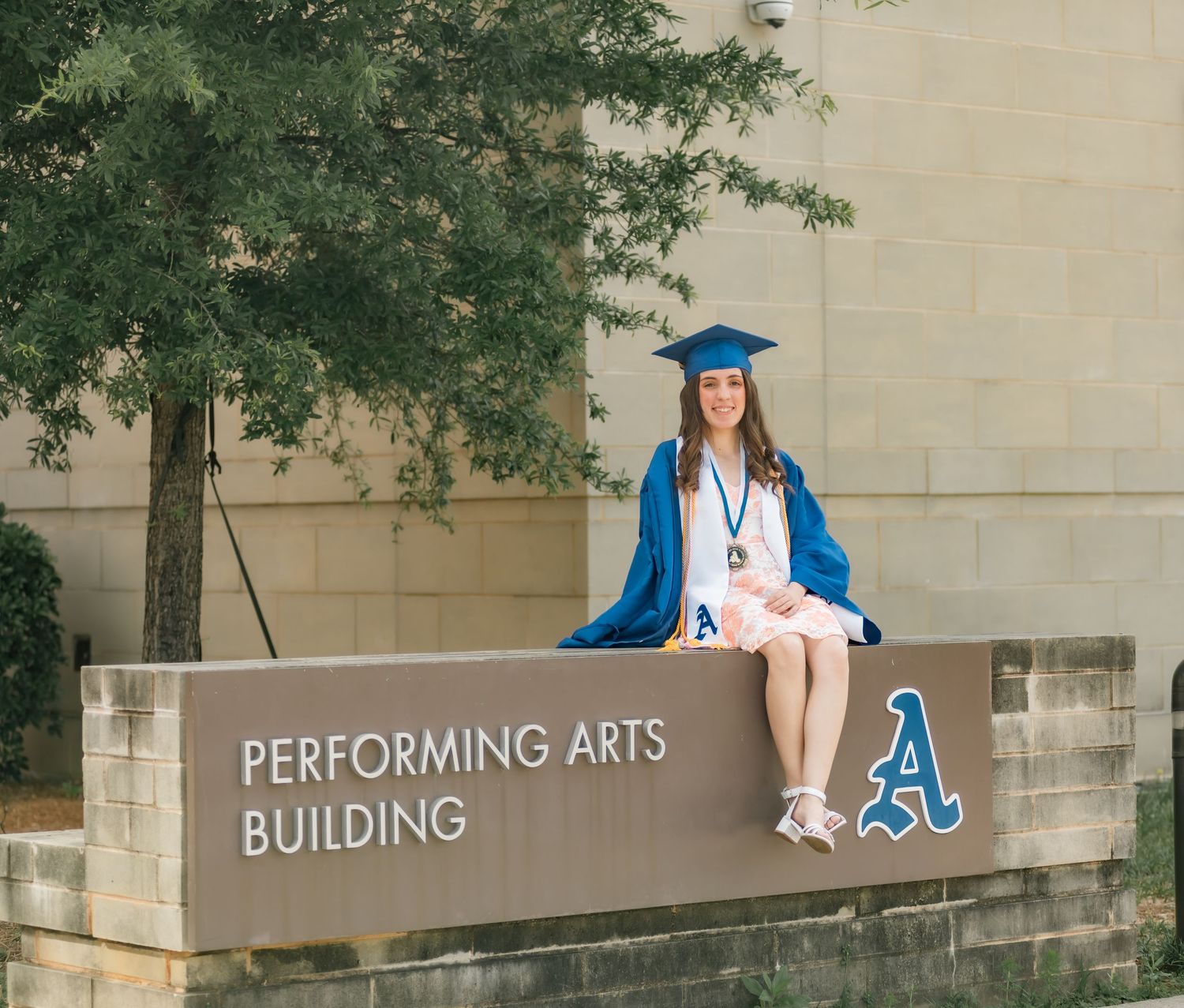 Graduate in blue cap and gown sits on Performing Arts Building sign at university campus.
