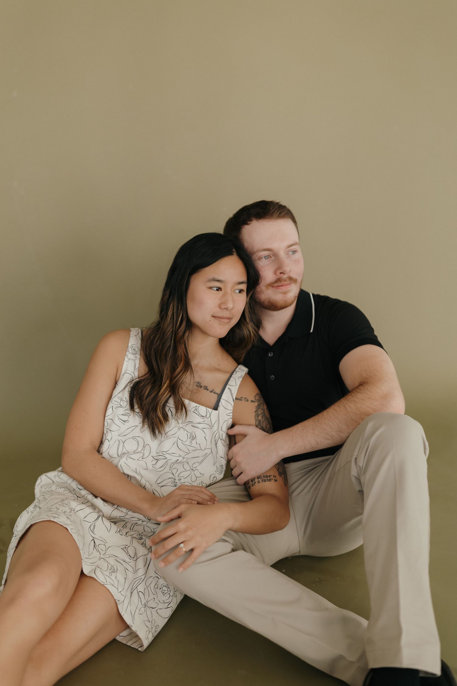 Intimate couple pose together against beige wall, wearing white dress and black shirt in minimalist portrait setting.