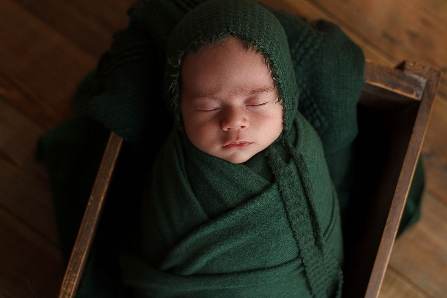 Newborn baby boy wrapped in emerald green with a matching bonnet sleeping in a wooden crate