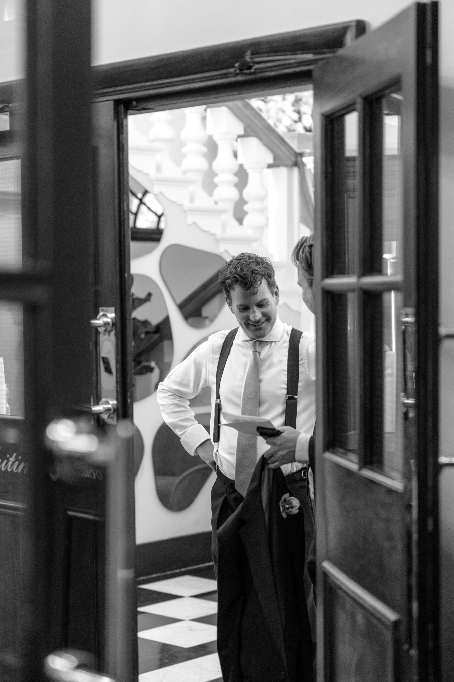 A black and white photo taken through a doorframe of someone adjusting their suspenders in a formal setting.