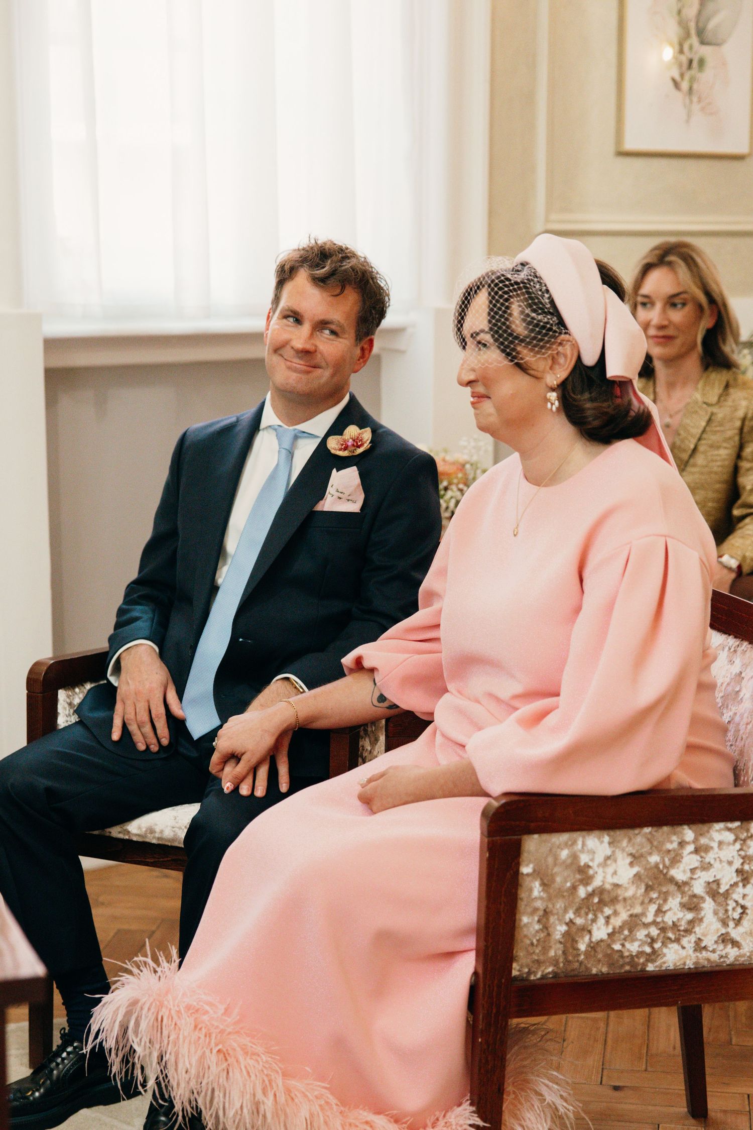 A wedding couple seated together, with the person in pink wearing a vintage-style dress and headpiece.