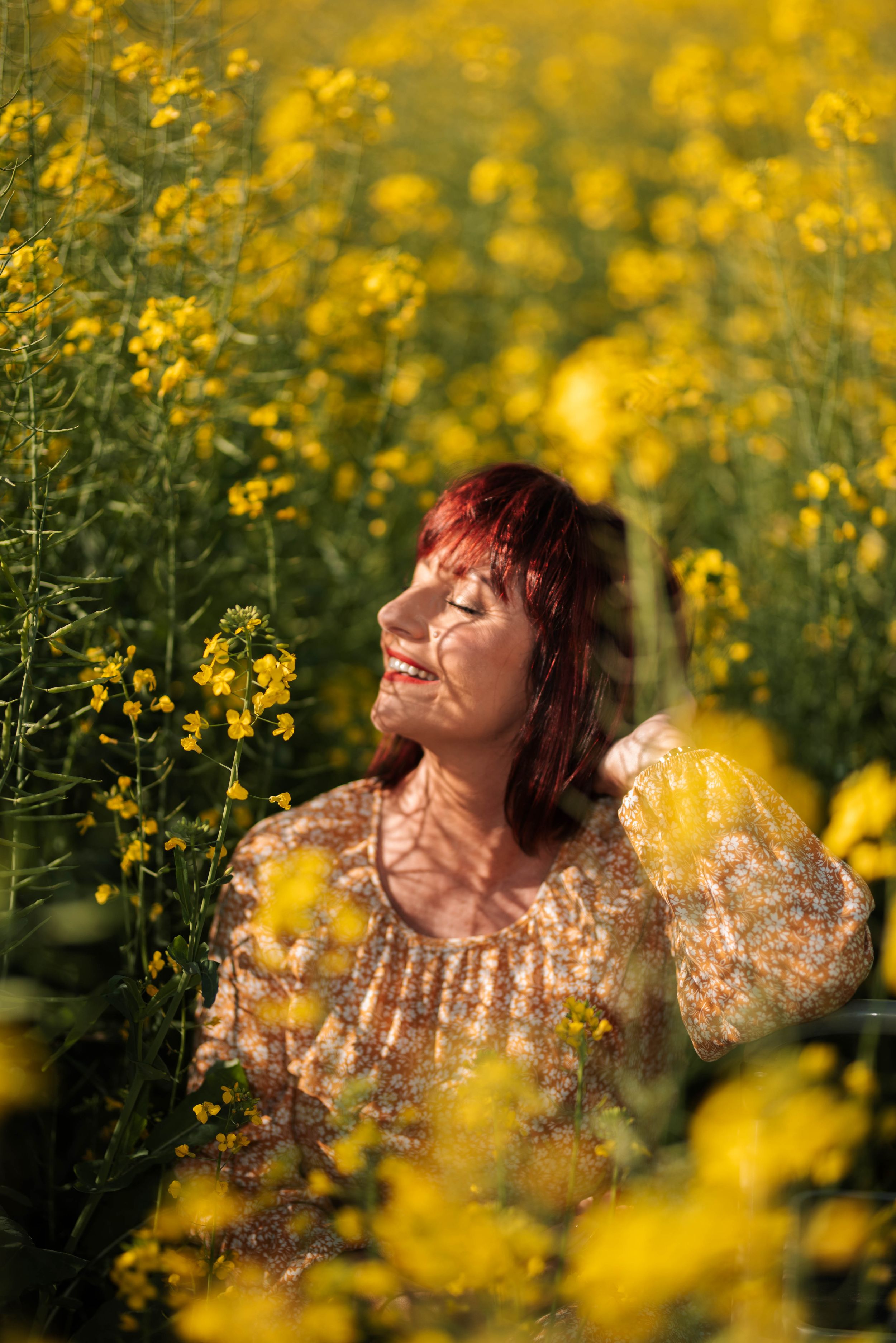Woman in floral dress poses joyfully in field of bright yellow wildflowers at sunset.