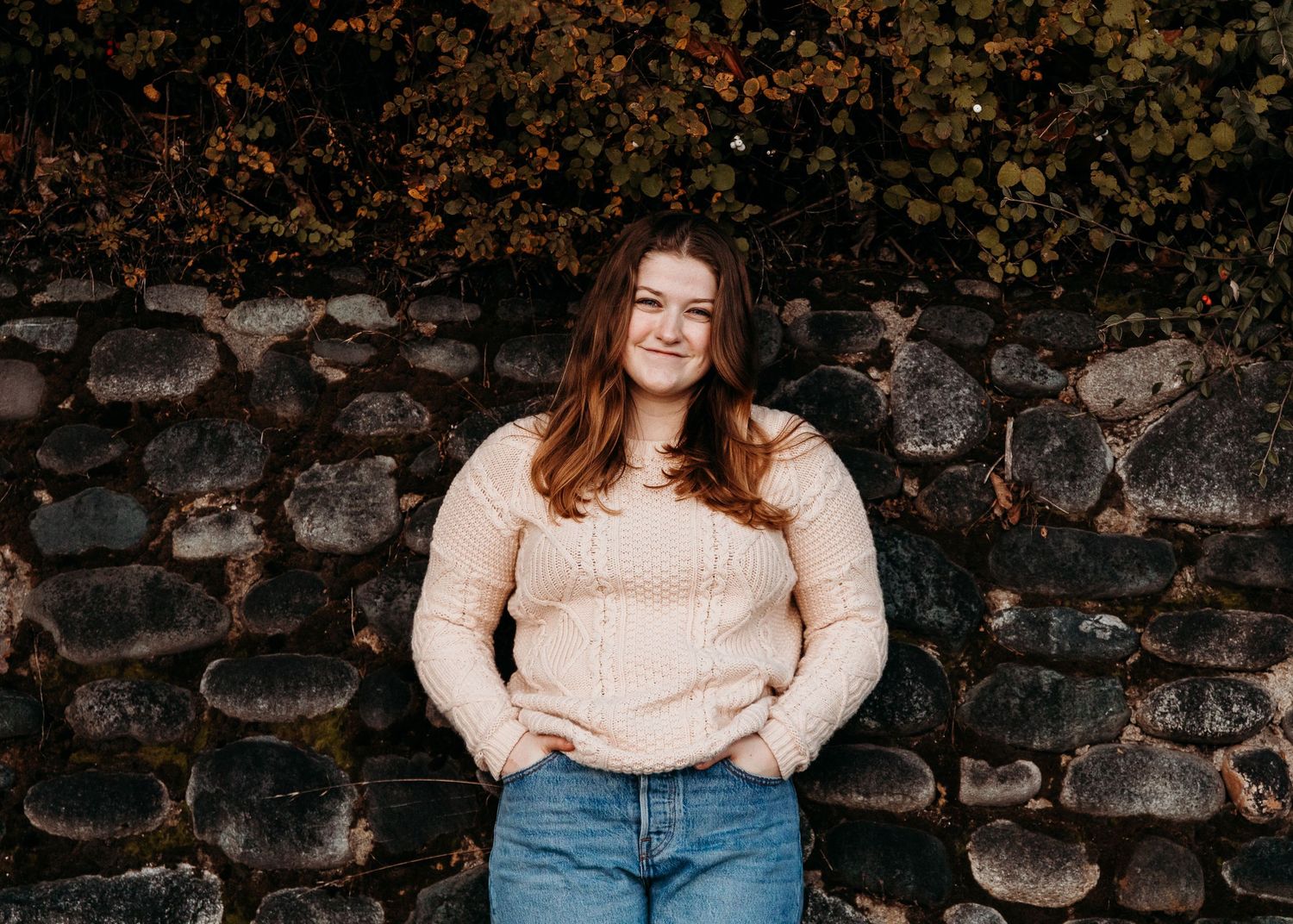 Person in beige sweater and blue jeans stands against rustic stone wall in autumn setting.