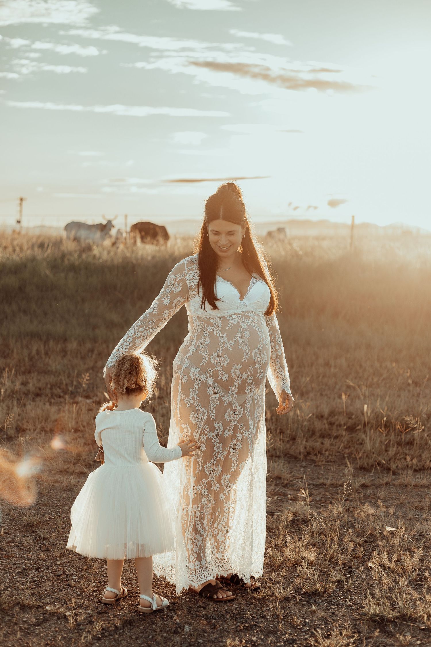 Mother in lace maternity dress walks with daughter in tulle dress through golden field at sunset.