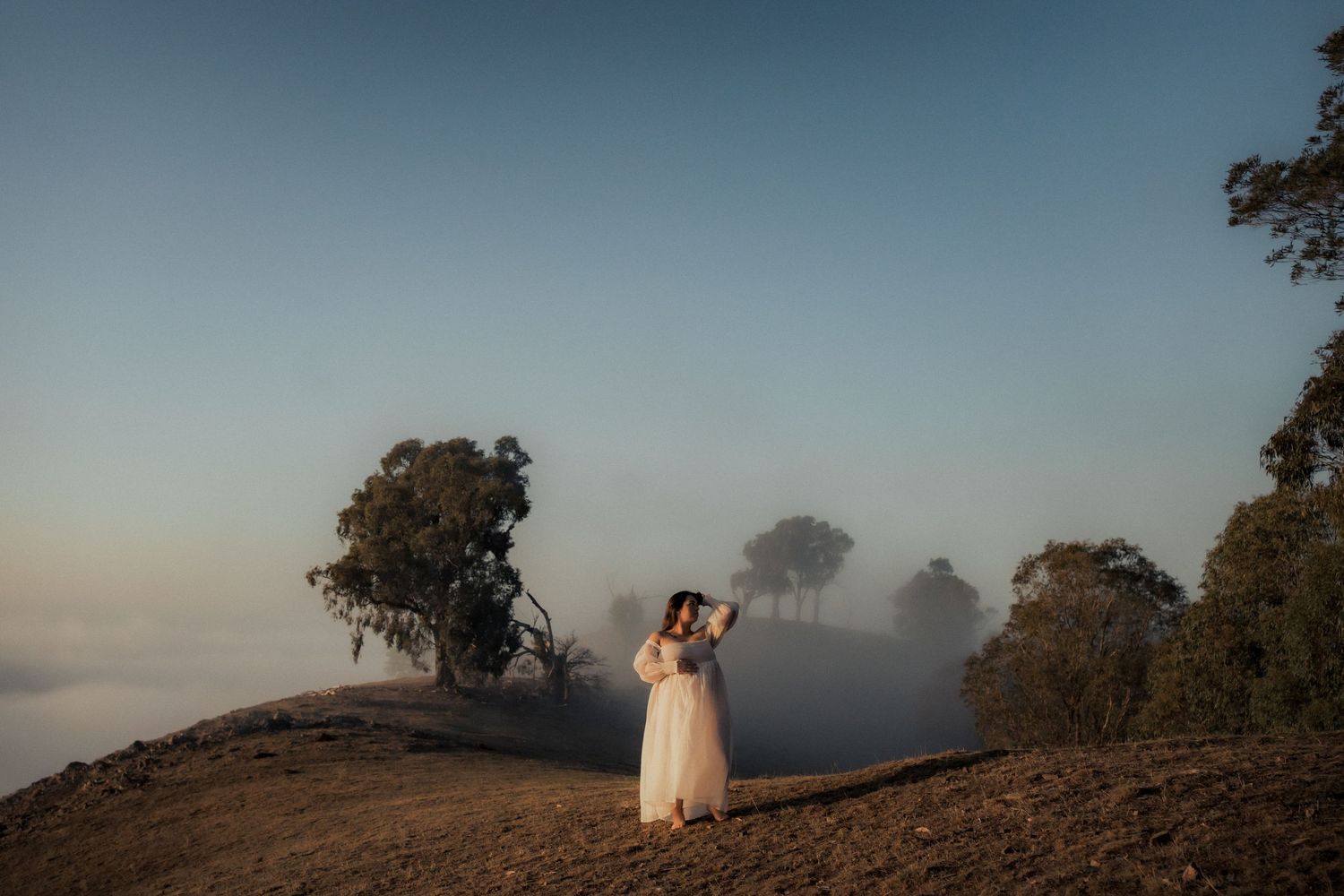 Dreamy hillside scene with person in white dress standing above foggy valley at sunset.