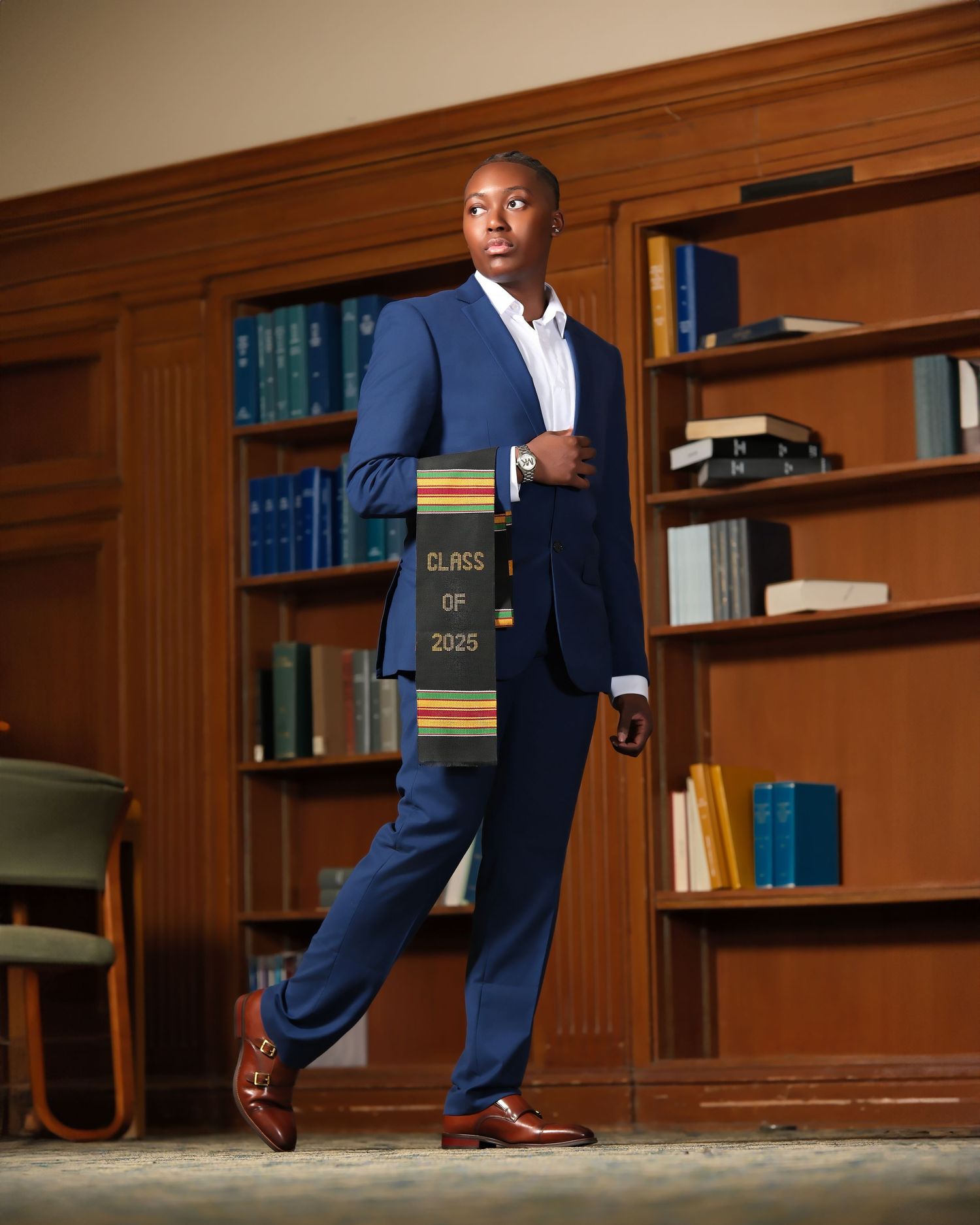A person in a navy blue suit walks confidently through a wood-paneled library while holding books.