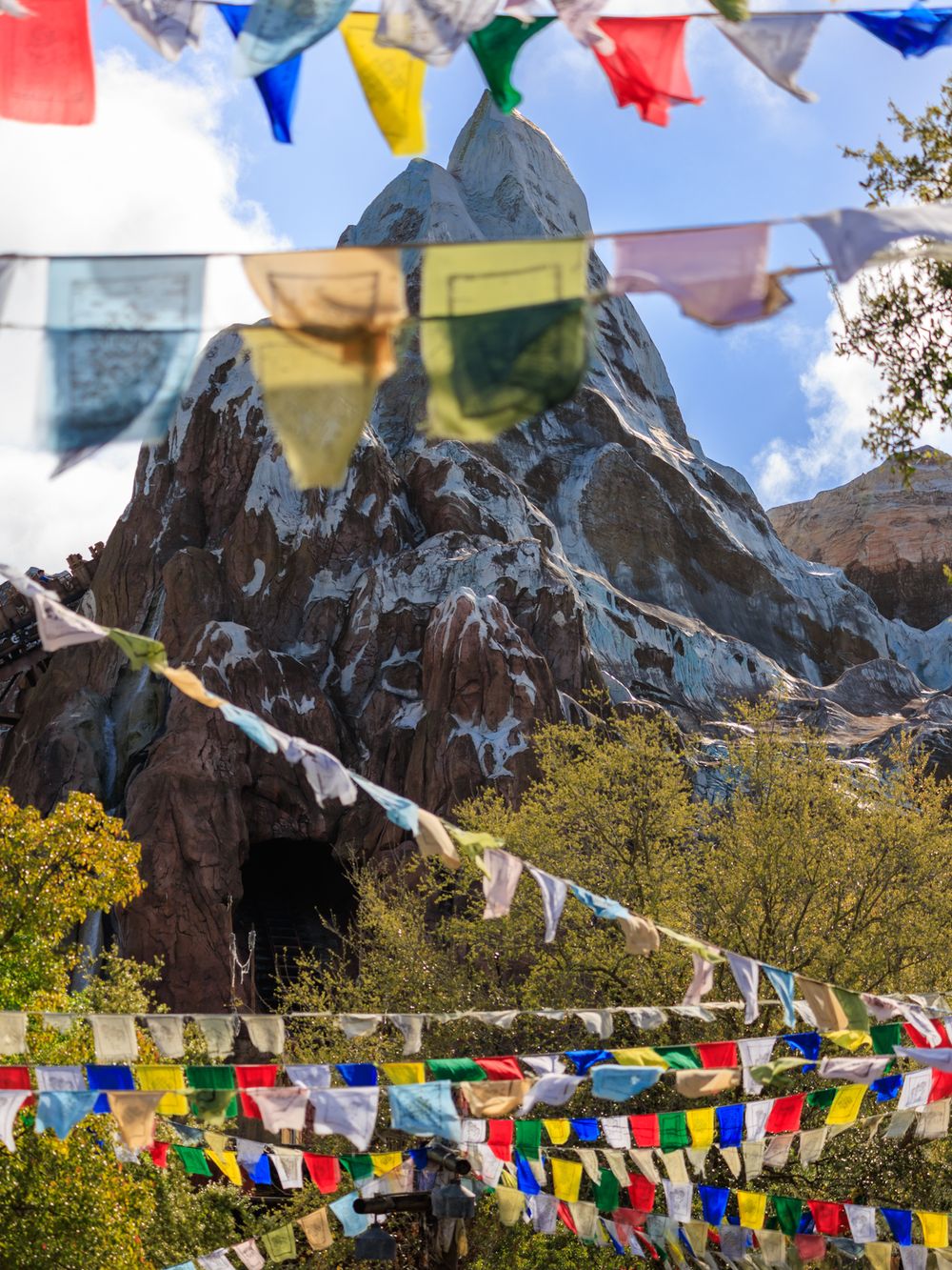 Colorful Tibetan prayer flags wave against a mountain backdrop with blue skies and evergreen trees.