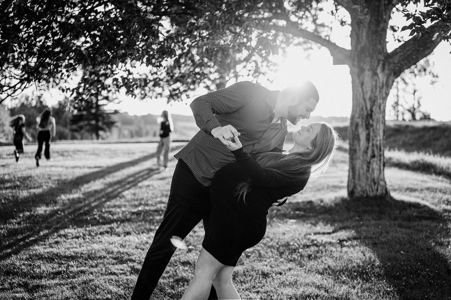 Photo romantique en noir et blanc d’un couple s’enlaçant sous un arbre au coucher du soleil.