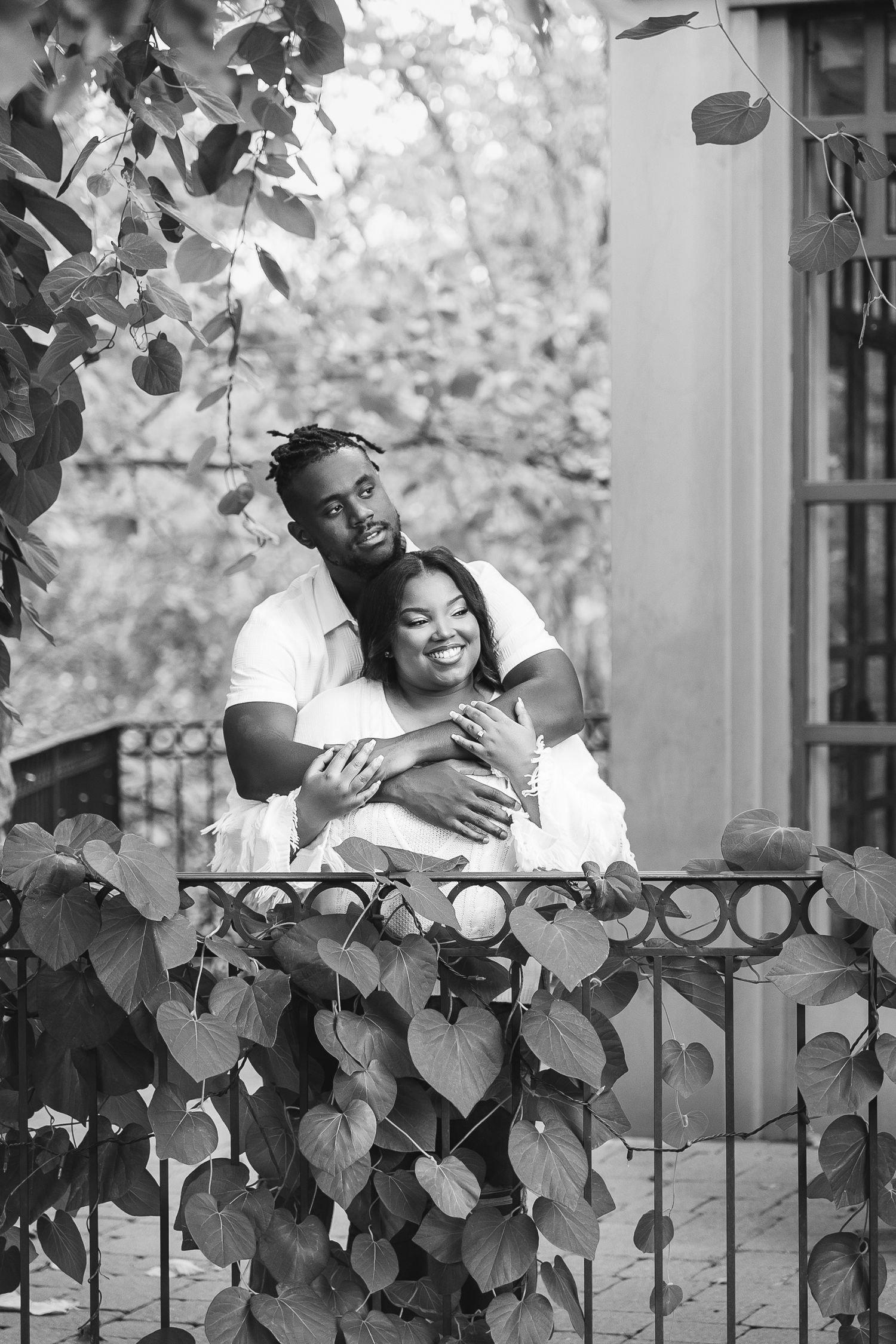 Black and white photo of happy couple embracing on vine-covered balcony with iron railing.