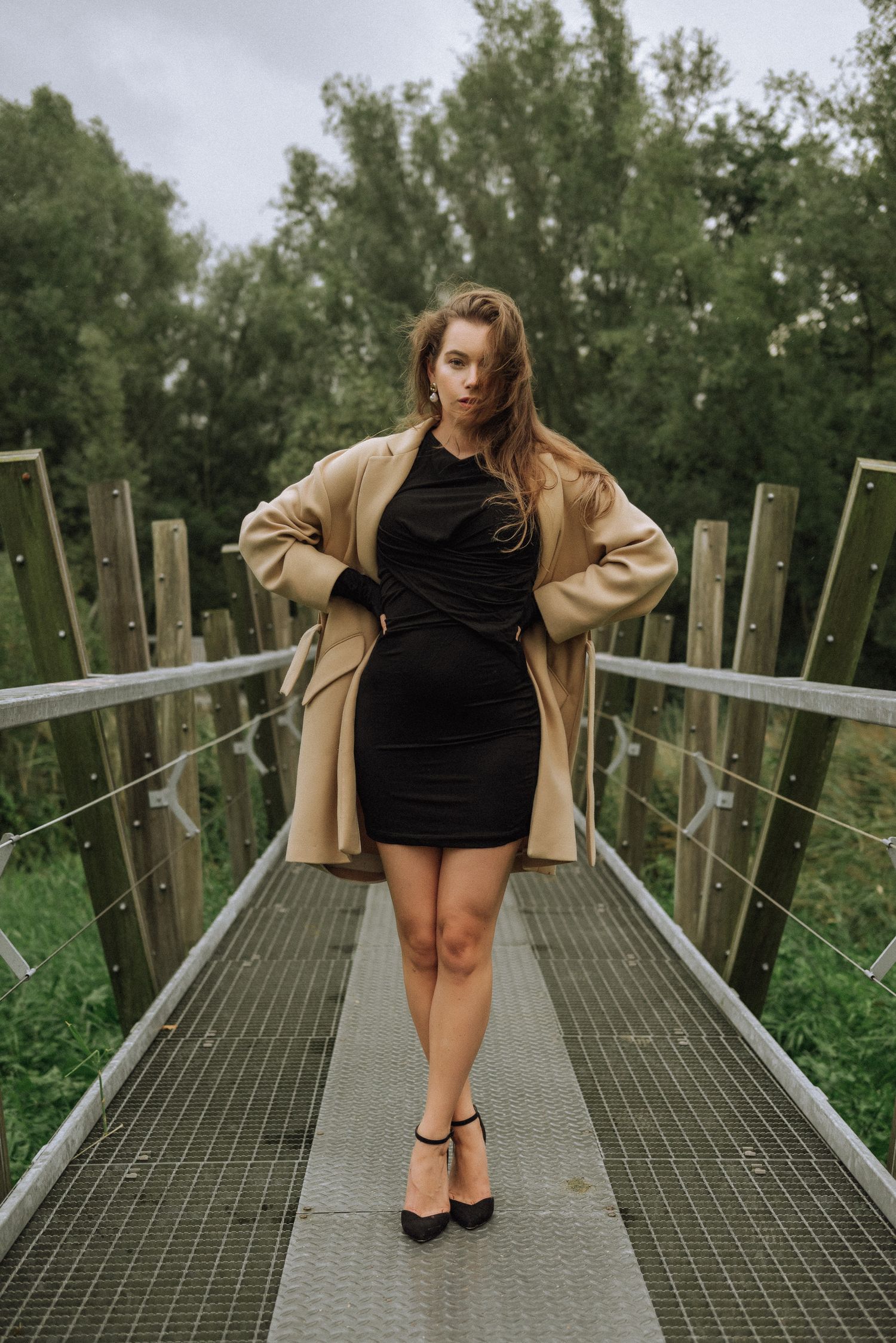 Figure in black dress and beige coat posing on metal walkway surrounded by greenery.