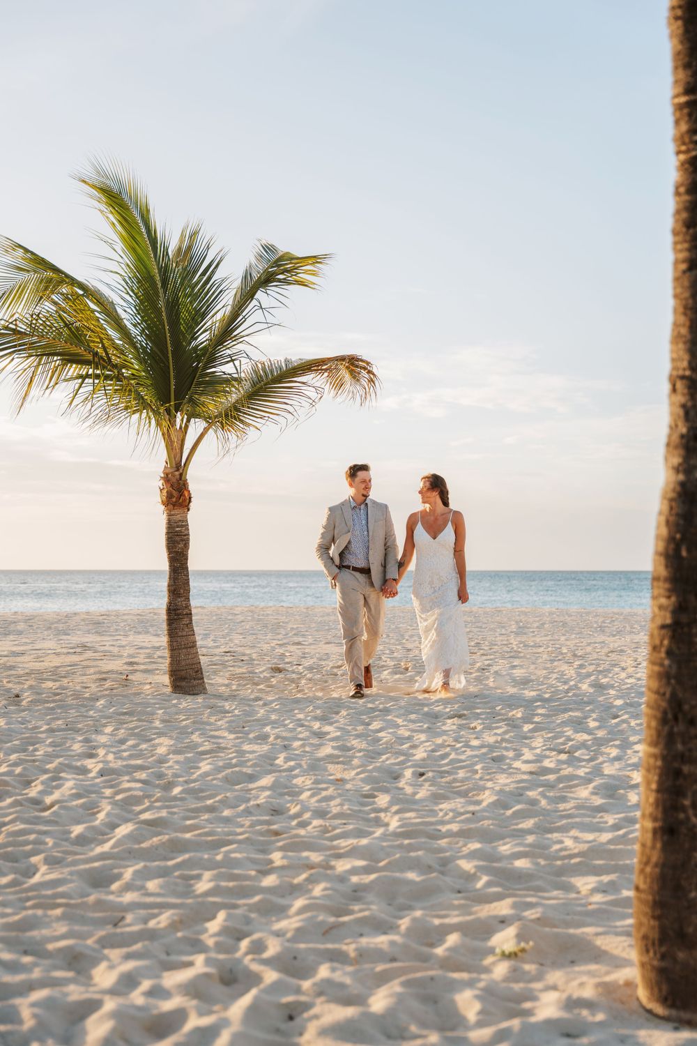 A couple walking hand in hand on a tropical beach with palm trees and soft white sand at sunset.