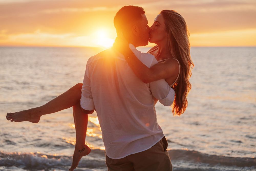 Silhouetted couple embraces on beach during golden sunset over calm ocean waters.