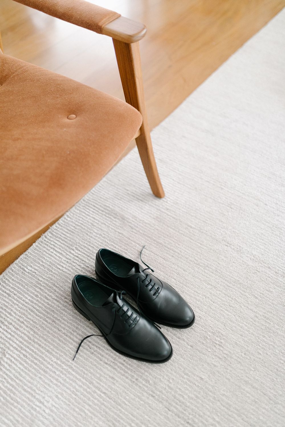 Black leather oxford shoes placed on a light carpet next to a tan velvet chair leg.