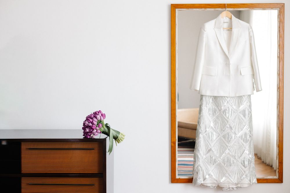 White wedding dress and jacket ensemble displayed on mirror with floral accent.