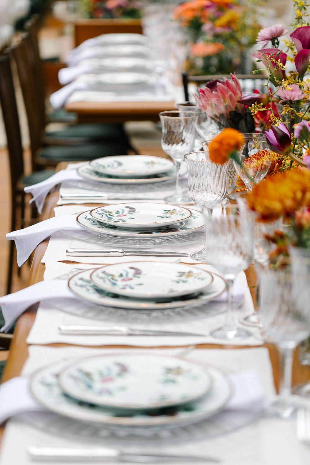 Formal table setting with patterned china plates and colorful floral centerpieces.
