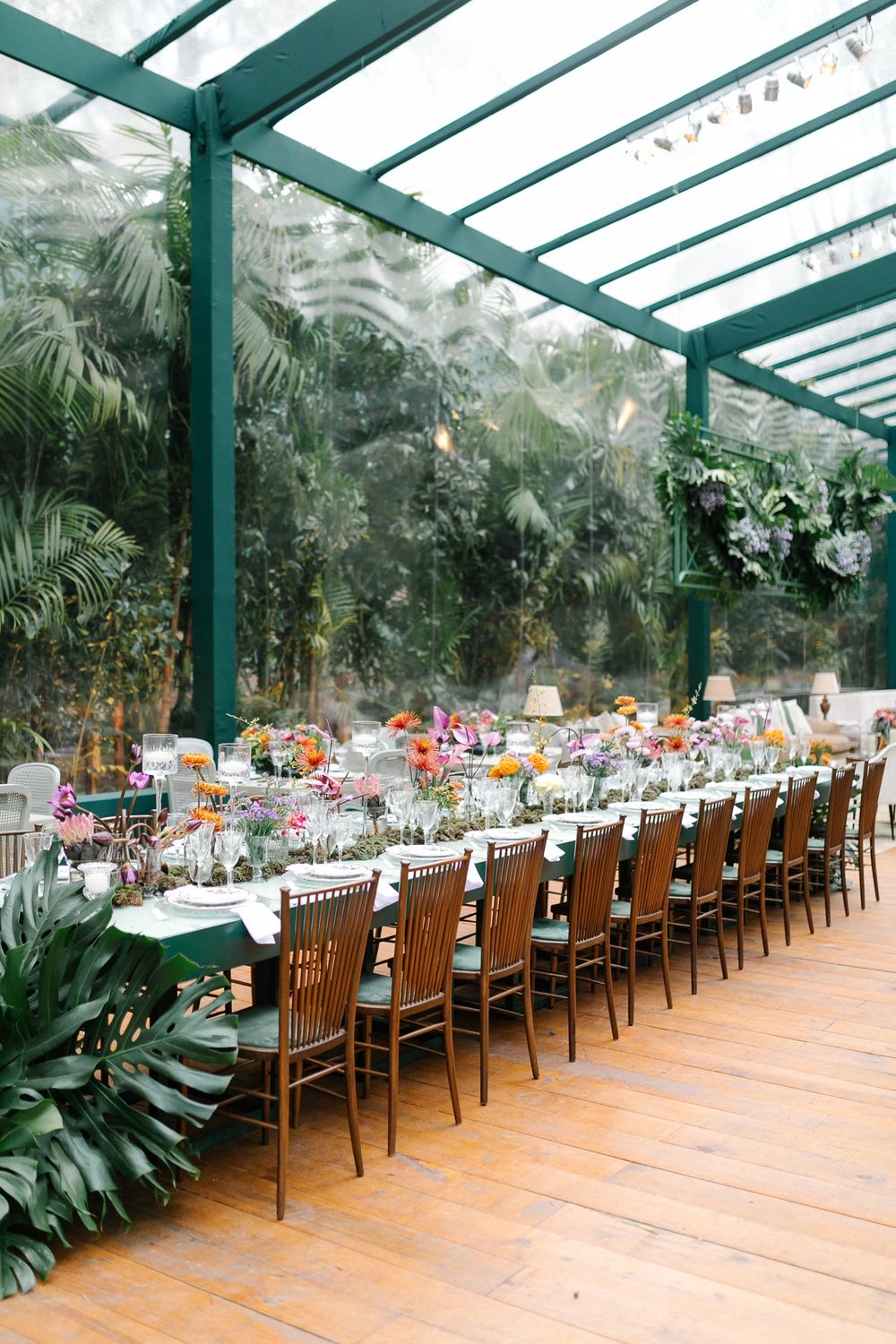Long dining table set in a greenhouse with lush tropical plants and hanging glass decor.