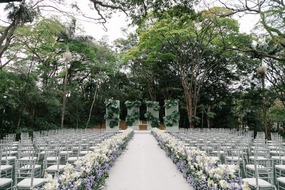 Outdoor wedding ceremony setup with white chairs and floral-lined aisle beneath tall trees.