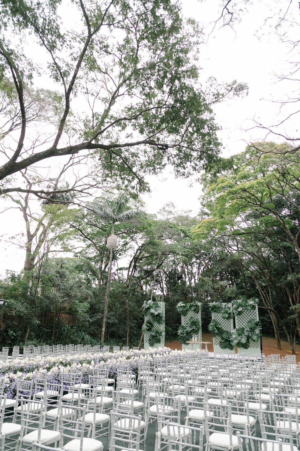 Wedding ceremony venue with rows of white chairs under a canopy of lush green trees.