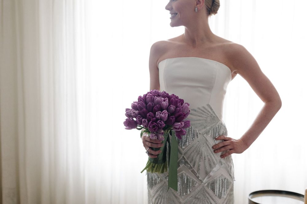 Elegant white strapless wedding dress with purple tulip bouquet against window light.