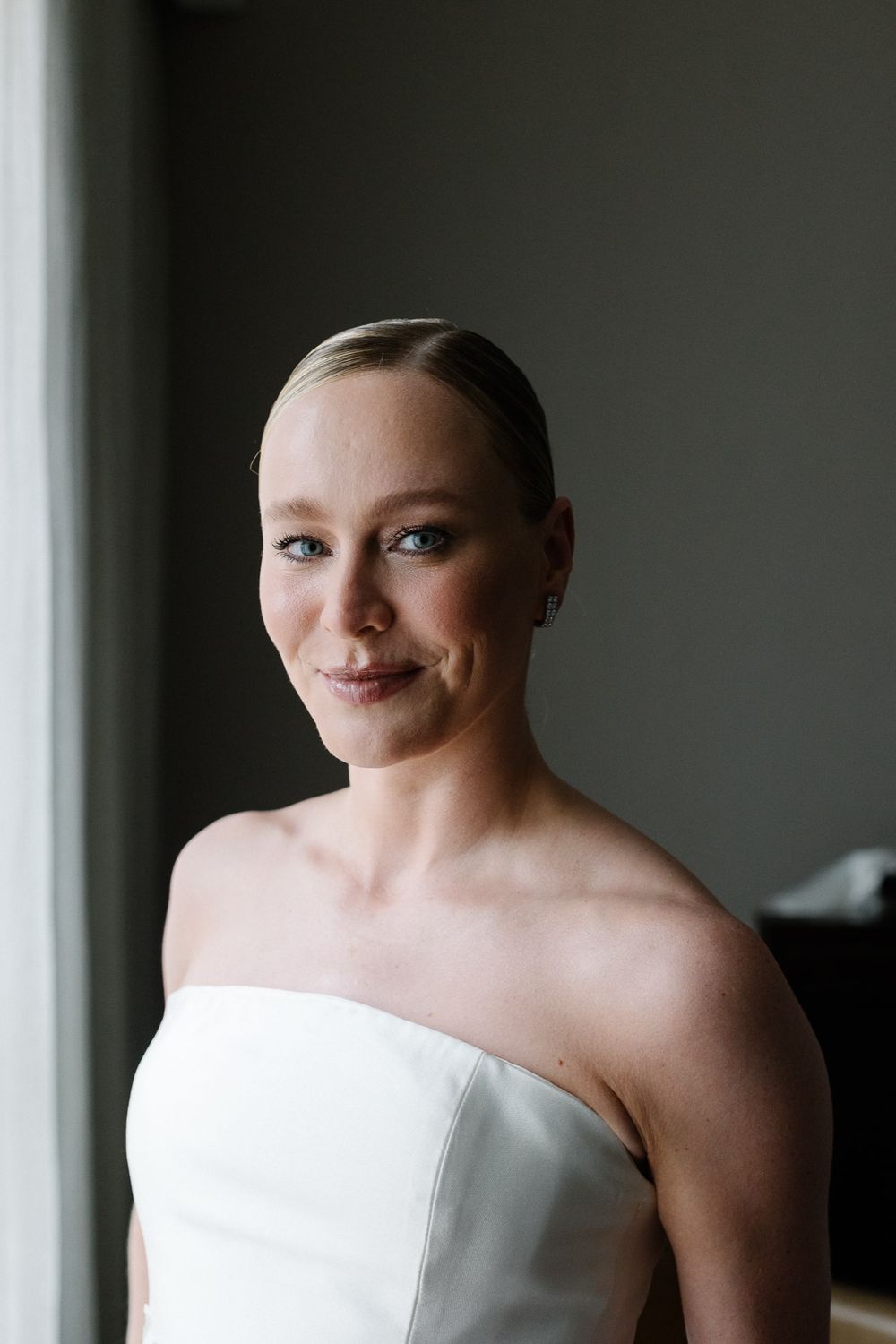 An elegant bridal portrait near a window in a classic white strapless gown.