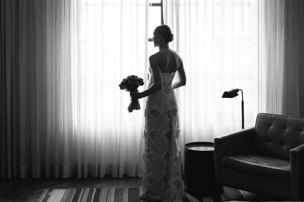 A silhouetted figure in a wedding dress stands elegantly by a window holding a bouquet, creating a dramatic backlit composition.