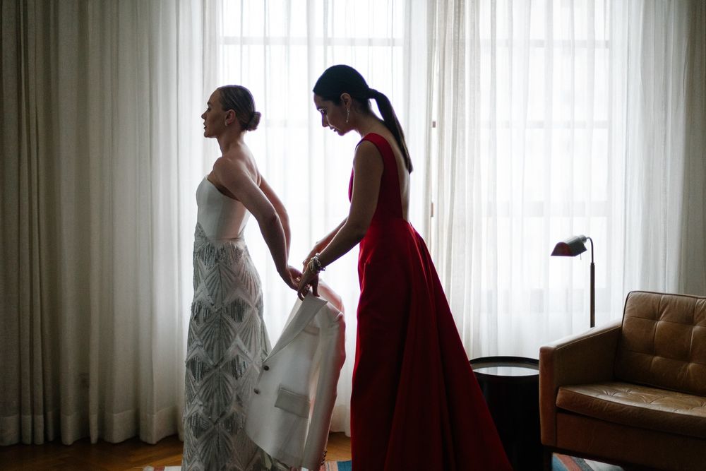 Two figures in silhouette by a window, one in a white dress and another in red helping with wedding preparations.