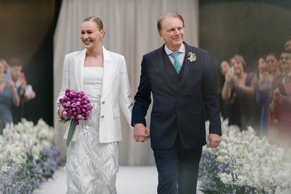 Newlyweds walking down an aisle lined with white flowers, bride holding purple bouquet.