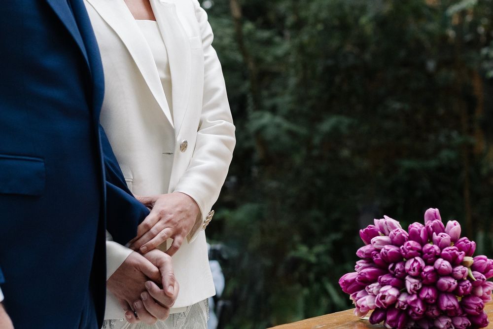 Close-up detail of joined hands with bright purple tulip bouquet and contrasting navy and white attire.