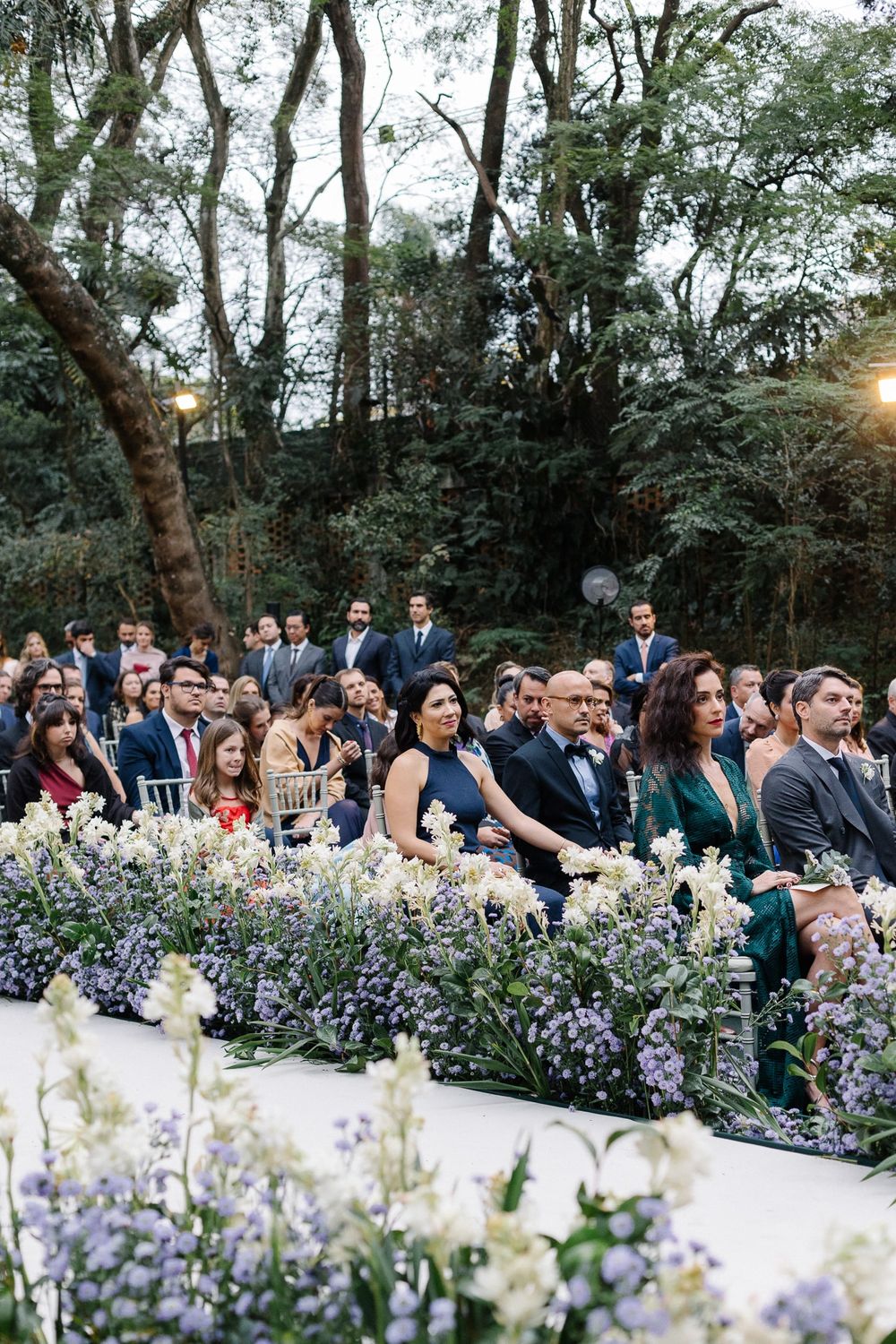 Outdoor wedding ceremony setup with guests seated among lavender and white floral arrangements in a wooded setting.