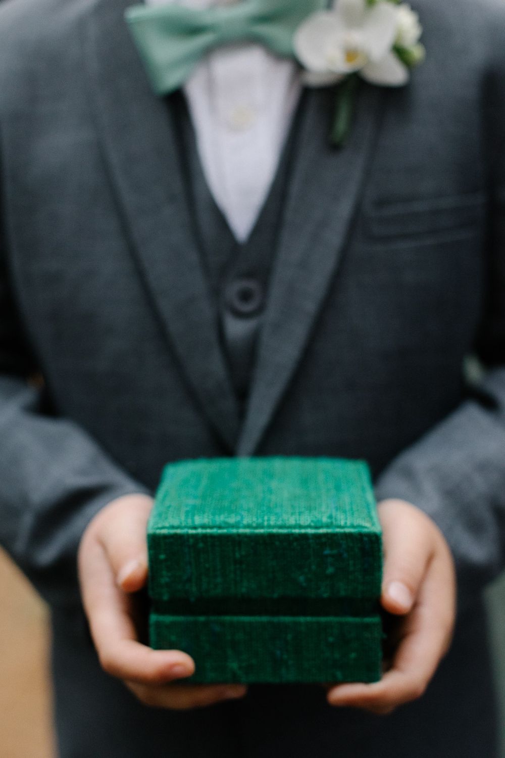 A person in a formal suit holding an emerald green velvet ring box with a bow tie.