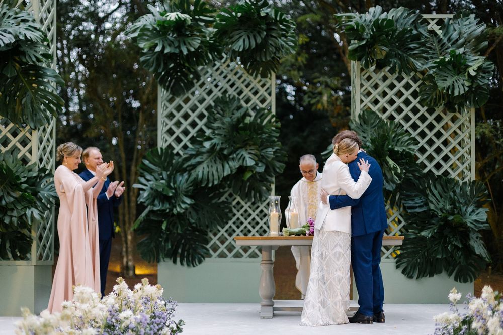 A wedding ceremony takes place in a tropical garden setting with white floral decor and lattice backdrop.