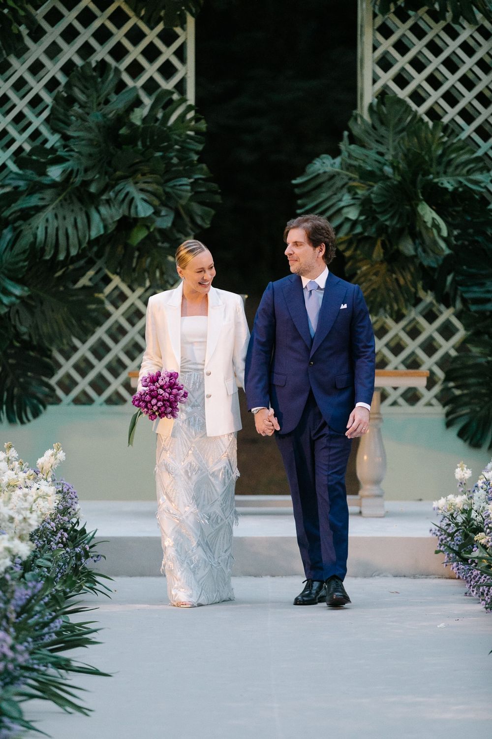 A couple walking together at their wedding with tropical decor and white lattice backdrop.