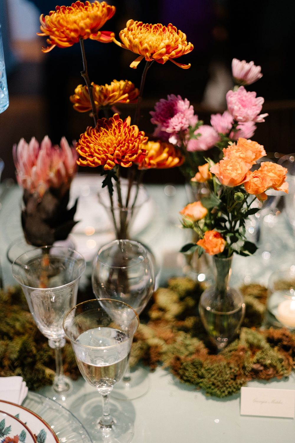 Elegant table setting with orange chrysanthemums, pink flowers and wine glasses arranged on moss-covered surface.