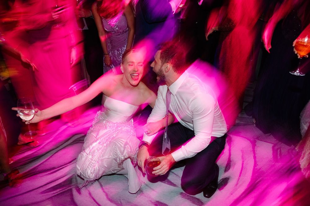 A couple dances energetically under vibrant pink lighting at a wedding reception.