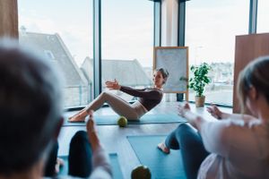 A fitness instructor demonstrates a seated boat pose during a yoga class in a bright studio space near windows.