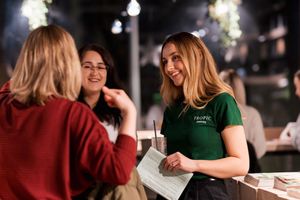 People having a conversation at an outdoor restaurant with one server in a green uniform shirt taking their order.