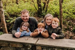 Family poses together while leaning on wooden table in a lush green forest setting.