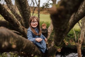 A young person wearing glasses and denim sits perched among the branches of a large tree on a cloudy day.