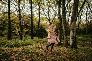 A person in a pink dress twirls playfully in a green autumnal forest setting surrounded by tall trees and fallen leaves.