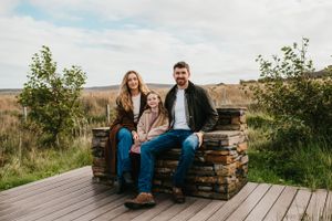 A family sits outdoors on a rustic brick wall on a wooden deck surrounded by prairie grasses and shrubs.
