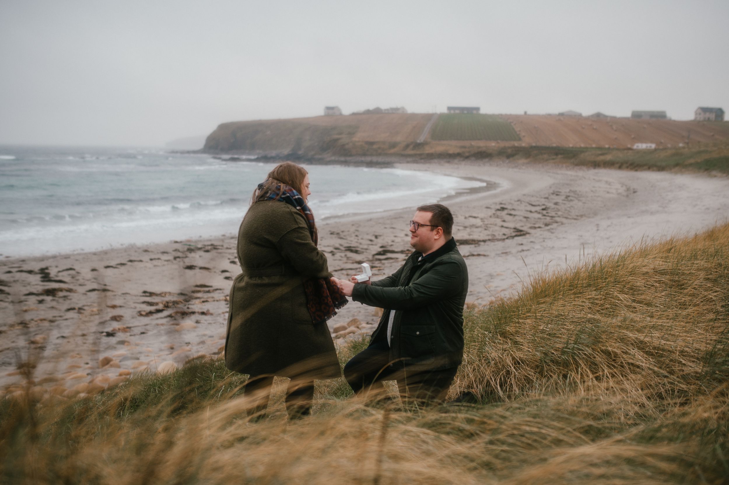 Two people share a quiet moment on a grassy cliff overlooking a misty beach with rocky coastline in the background.
