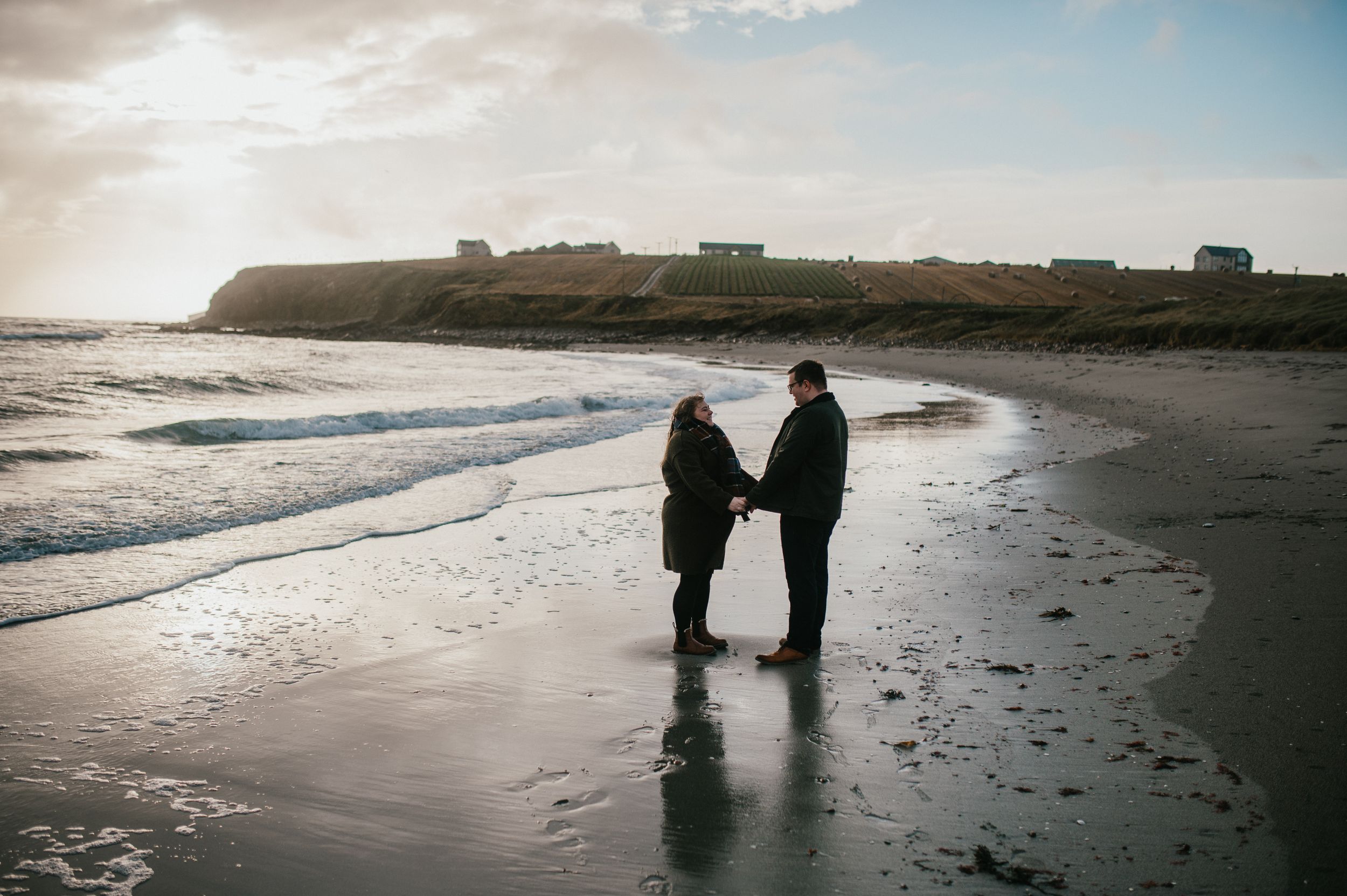Two silhouetted figures stand together on a beach at sunset with dramatic cliffs in the background.